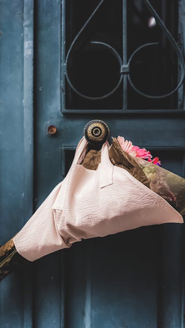 A person wearing a brown glove is holding a bouquet of fresh pink flowers wrapped in pink paper, about to deliver it through a small, round door with an ornate black metal grille. The background featu