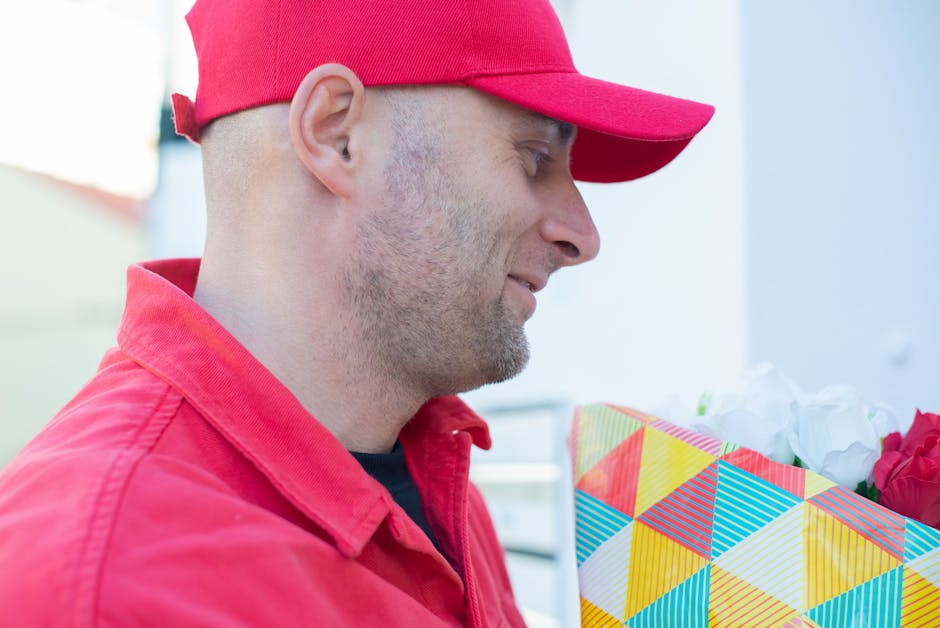 A man dressed in a red cap and red shirt is holding a colorful floral gift bag with a geometric pattern, filled with white and pink flowers. The background appears to be a light-colored, outdoor setti